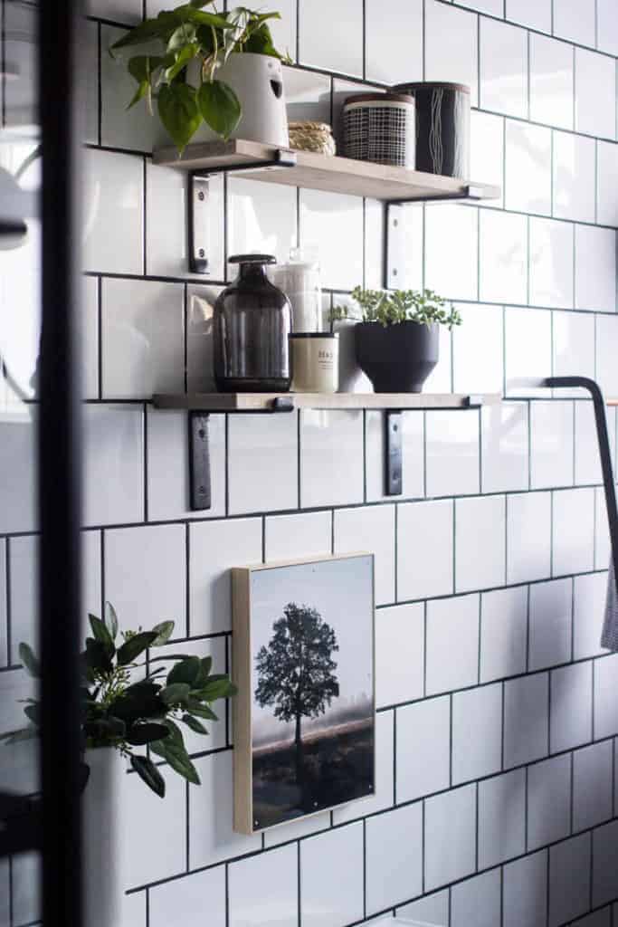 White square tile wall with dark grout and two floating shelves with black brackets drilled into tile with various vases and plants as decor