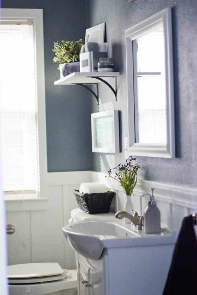 Blue walls in bathroom with white board and batten on the bottom, white shelf hanging above toilet, mirror with white frame