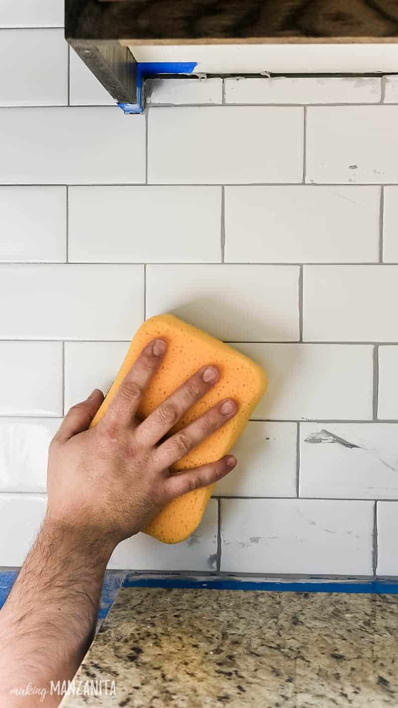 Wiping off excess gray grout on white subway tile backsplash in kitchen with a wet yellow sponge