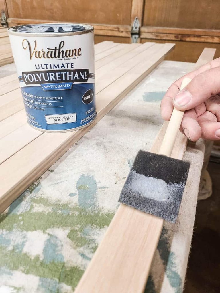 Can of matte polyurethane on top of slat of wood and man brushing matte polyurethane using foam brush onto the wood slat to seal the wood