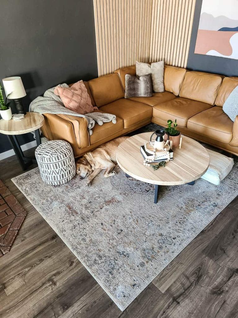 Above image of seating area in modern boho living room with light brown leather sectional, wood slat accent on the wall, tapestry wall art and Lab sleeping between sectional and coffee table.