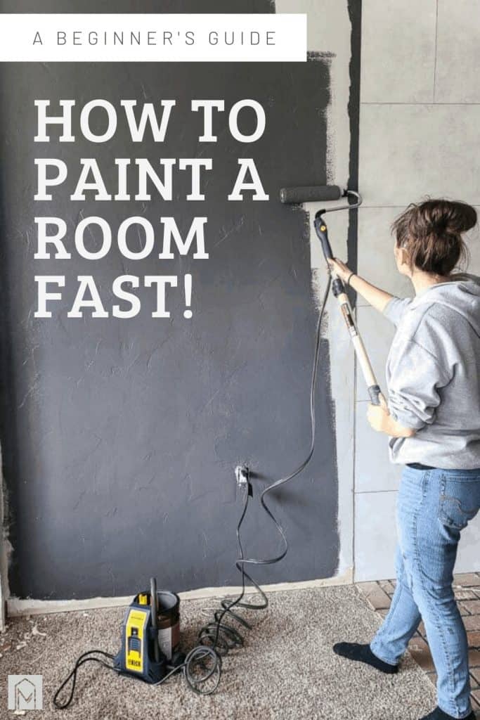 A woman wearing jeans and a gray sweatshirts is pictured painting a wall with gray paint using a power paint roller.